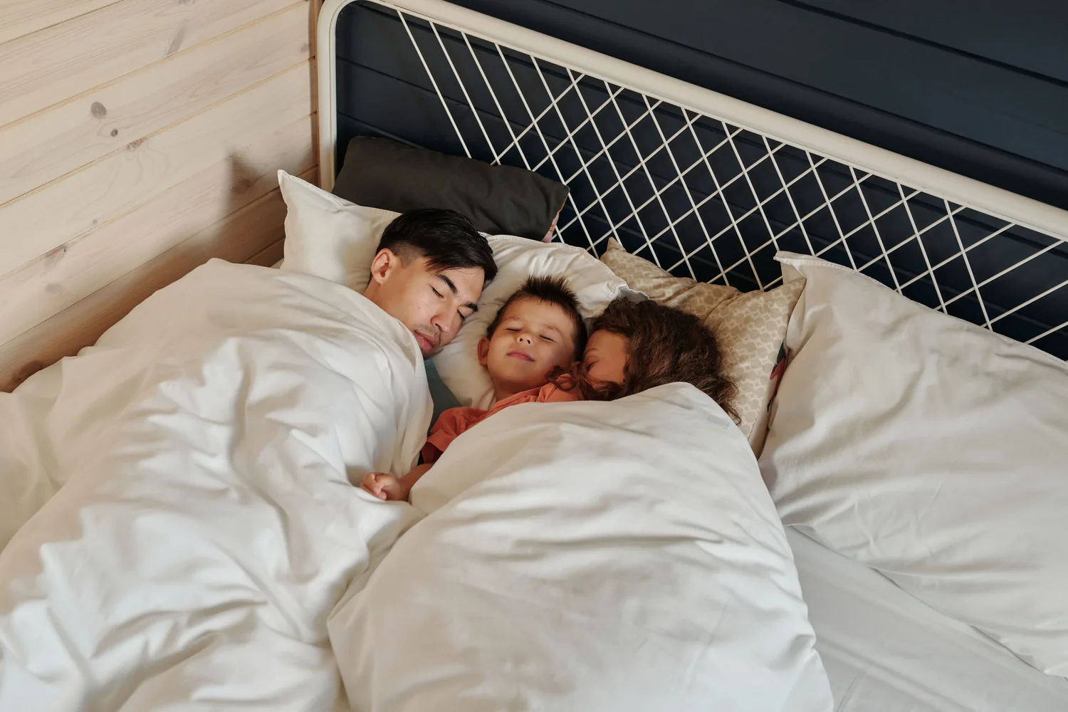 Papá dormido en la cama abrazando a sus dos hijos pequeños bajo un edredón blanco
